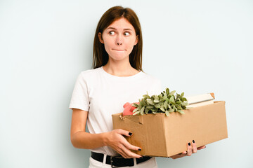 Young English woman making a move while picking up a box full of things isolated on blue background confused, feels doubtful and unsure.