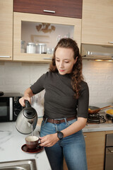 Young woman standing at kitchen counter and puring herself cup of hot tea