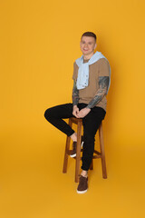 Fototapeta premium Smiling young man with tattoos sitting on stool against yellow background