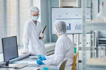 Two chemists in lab coats talking to each other while working with statistics of epidemic in the laboratory