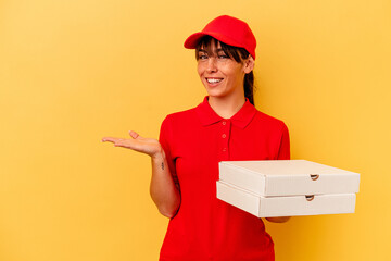 Young delivery woman holding pizzas isolated on yellow background showing a copy space on a palm and holding another hand on waist.