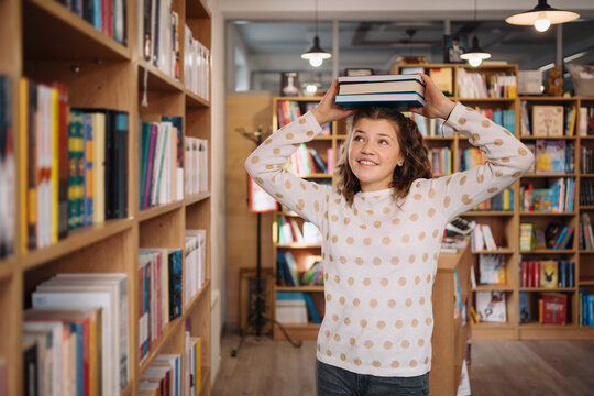 Teen girl among a pile of books. A young girl holding books on head with shelves in the background. She is surrounded by stacks of books. Book day.