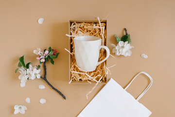 An open gift box with peach-colored paper shavings containing a creative trendy mug with a geometric pattern, a white paper bag and apple flowers on a beige background. Flat lat, top viw