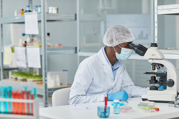 African young scientist in mask examining sample of virus with microscope while sitting at the...