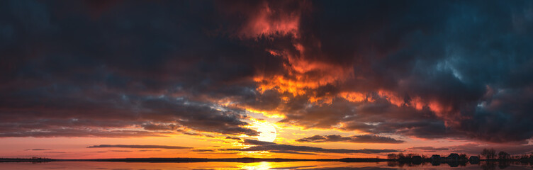 A bright sunset with a large yellow sun reflected in the water. Sunrise at the edge of the village. The big sun goes over the horizon reflecting the light in the lake