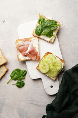 White toasts with cream cheese, ham, cucumbers and spinach on a white marble board