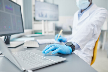 Close-up of scientist in protective gloves typing on laptop at the table and making notes in card...