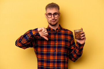 Young caucasian man holding a almond jar isolated on yellow background showing a dislike gesture,...