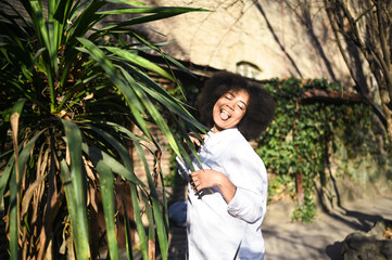 Fashion close up stylish portrait of attractive young natural beauty African American woman with afro hair in white shirt posing outdoors with green palm. Happy lady laugh with perfect smile and teeth