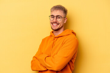 Young caucasian man isolated on yellow background who feels confident, crossing arms with determination. - obrazy, fototapety, plakaty