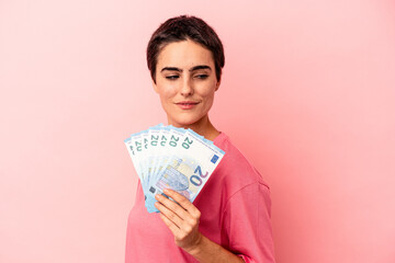 Young caucasian woman holding banknotes isolated on pink background looks aside smiling, cheerful and pleasant.