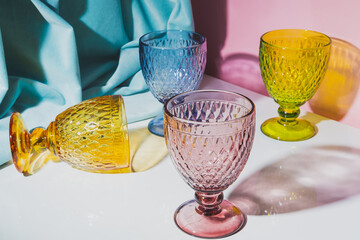 Four stylish colorful faceted glasses with shadows on a white table with a pink background and turquoise drapery. - 479564163