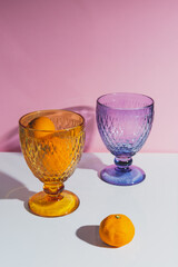Stylish orange and blue faceted glasses on a white table with tangerines with pink background.