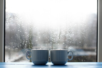 pair of mugs with frothy cappuccino against the background of a window with drops after the rain. welcome spring with warming coffee