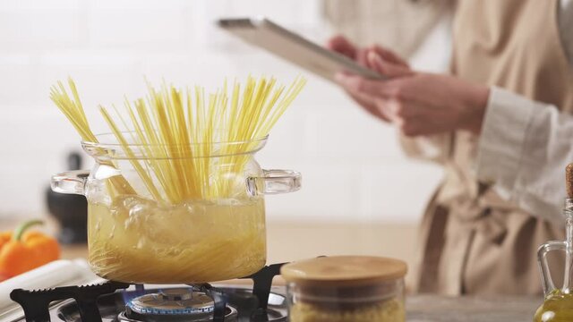 Hands hold a tablet in the kitchen. Passionate about the tablet while cooking spaghetti on the stove in a transparent bowl. Cooking macaroni in the kitchen stove boiling water boiling water in a glass