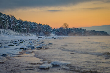 deep snow over rocks at the beach in cold winter sunset by the sea with pine trees and spruce
