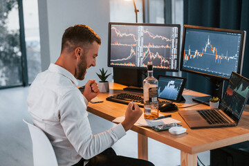 With alcohol. Young businessman in formal clothes is in office with multiple screens