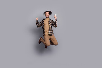 Photo of excited lucky guy dressed checkered shirt jumping high typing modern device isolated grey...