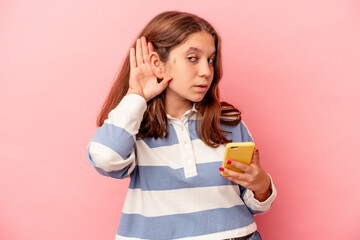 Little caucasian girl holding mobile phone isolated on pink background trying to listening a gossip.