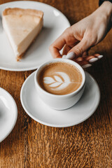 Woman is holding a cup of coffee on a wooden table near a cheesecake.