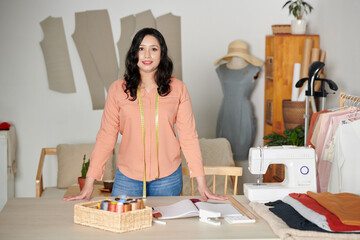 Portrait of smiling seamstress standing at table in her home workshop