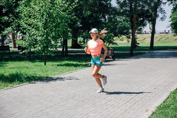 running fast smiling young sporty woman with watch in park in pink t-shirt, green shorts , white cap and trainers on background of green trees and grass in front of bench and inscription on flowerbe
