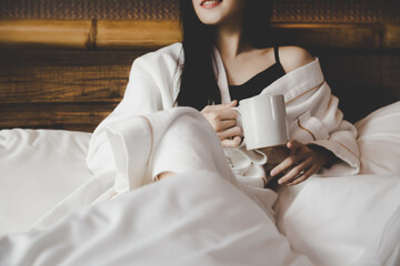 Young beautiful girl wearing bathrobe drinking coffee on bed in cozy bedroom in the morning rituals...