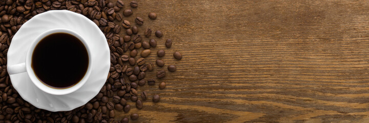 White cup of black coffee with brown beans on dark wooden table background. Closeup. Wide banner. Empty place for text. Top down view.