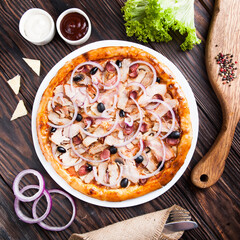 Meat pizza with onions and slices of pork and grated cheese lies on a white plate on a wooden table