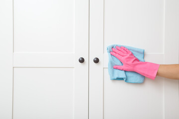 Young adult woman hand in pink rubber protective glove using blue dry rag and wiping white wooden doors of wardrobe. Closeup. Regular cleanup at home. Front view.