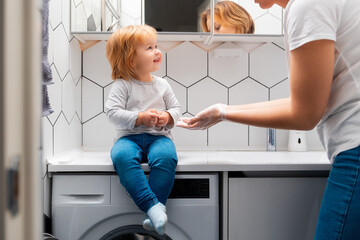 A little girl is sitting on the countertop in the bathroom and learns from a teacher how to wash...