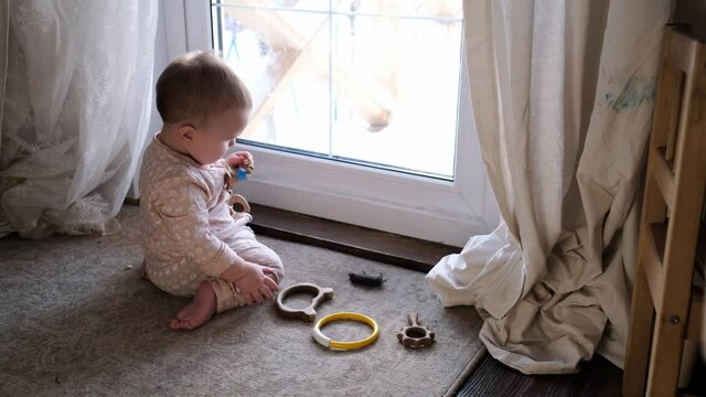 A cute plump Caucasian baby 10 months old is gnawing a toy on the floor near the door, window. Safe home space, real interior