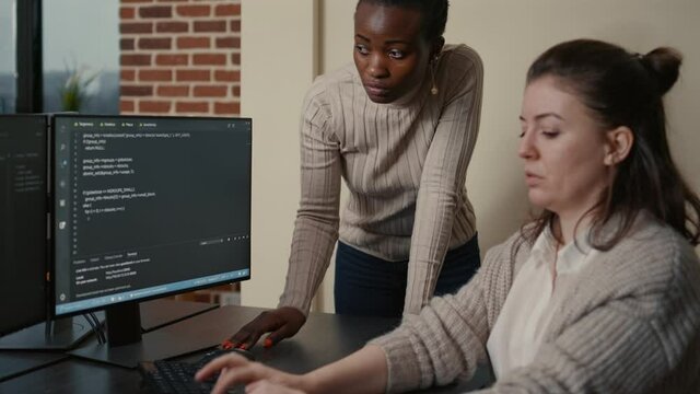 Junior developer writing source code under senior dev supervision sitting at desk with multiple screens showing programming interface. Programer writing algorithm with the help of colleague.