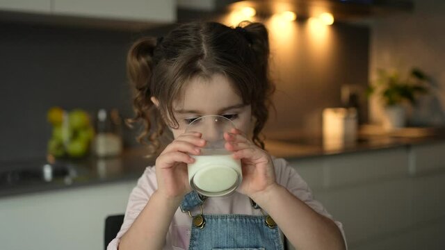 Little child drinking milk from a glass cup in her kitchen. Milk source of calcium for children.