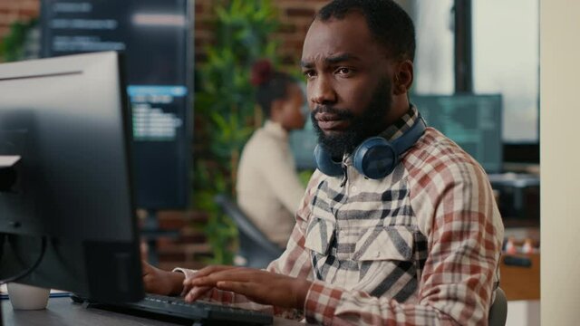 Portrait of focused african american programer wearing wireless headphones working looking at computer screen while typing. Software developer writing code in it agency office.