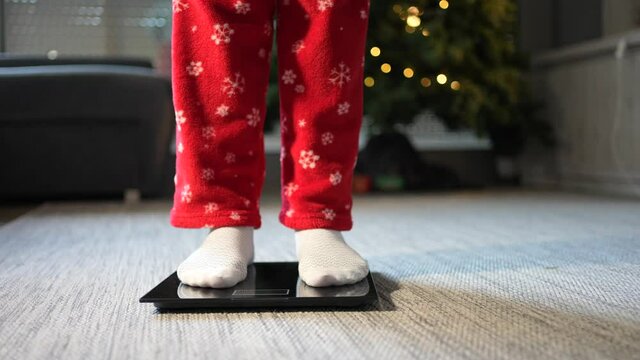 View of female feet climbing on electronic scale to control weight after Christmas holidays with Christmas tree in the background. Concept of bad nuricion at Christmas.