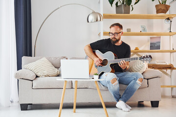 Using laptop. Man in casual clothes and with acoustic guitar is indoors