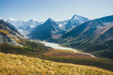 Wonderful alpine landscape with mountain lake and mountain river in valley with forest in autumn colors on background of snowy mountains silhouettes under blue sky. Beautiful mountain valley in autumn