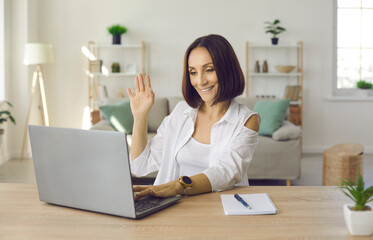 Friendly senior business woman waving in front of laptop webcam welcoming online conference participants. Woman enjoys virtual business communication and online negotiations sitting in her home office