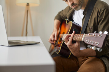 Beginner musician learning to play acoustic guitar. Pensive mature man plucking strings of his instrument while watching online rock or country music chords tutorial on modern laptop computer at home