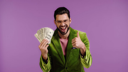 cheerful man in green jacket and glasses holding dollars and showing thumb up isolated on purple
