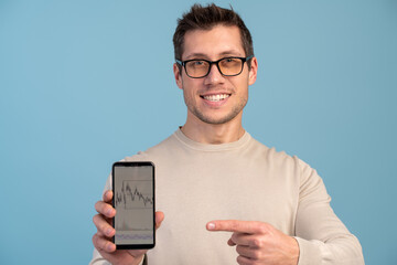 Portrait of satisfied brunette man with beard pointing at cellphone and smiling at camera, recommending gadget or mobile application. Indoor studio shot isolated on blue background