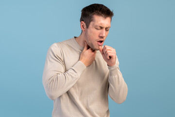 Feeling bad. Young caucasian man suffers pain in throat and coughing due a virus or infection while standing isolated over blue wall. Stock photo