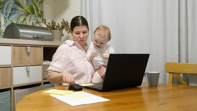 Woman holding a baby and working on her laptop, taking notes on paper. Young mother working with bills or have part-time job online. Little girl play with soft spoon