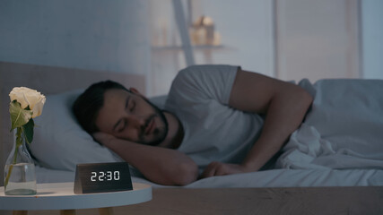 Clock and flower in vase near blurred man sleeping at home.