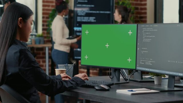 Asian programer writing code in front of computer with green screen chroma key mockup in software development agency. Coder computing big data with team colleagues working online cloud computing.