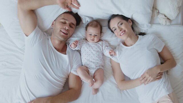 Top view portrait of loving and joyful young caucasian parents, mother, father and newborn baby daughter lying on white bed sheet and smile together and look at the camera. Happy young family concept
