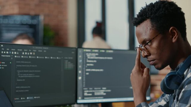 Portrait of focused software developer writing code arranging glasses sitting at desk in data room. System engineer programming user interface in busy artificial intelligence developing agency.