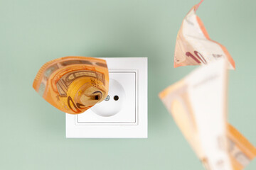 Euro money paper banknotes fall into a white electric socket on light green wall. Increasing cost...