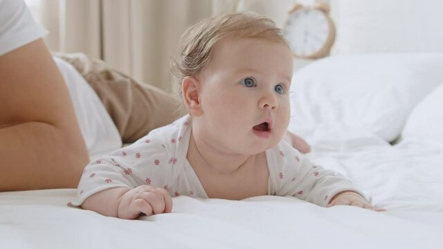 Cute sweet baby newborn with blue eyes lying on white bed sheet on her tummy expressive looks, moving her arms. Third month closeup portrait. Father gently strokes and kiss the child's head